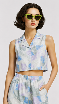 A model wearing the EVERSHE Chromaflora Ocean Lilac floral collared crop-top and wide-leg trouser co-ord set in a light blue, purple, and white watercolor print, shows close up view of the top, styled with lime green sunglasses, a green handbag, and green heels against a light gray studio background.