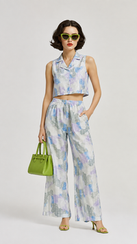 A model wearing the EVERSHE Chromaflora Ocean Lilac floral collared crop-top and wide-leg trouser co-ord set in a light blue, purple, and white watercolor print, shows the full front view of the dress, styled with lime green sunglasses, a green handbag, and green heels against a light gray studio background.