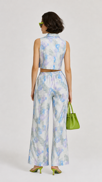 A model wearing the EVERSHE Chromaflora Ocean Lilac floral collared crop-top and wide-leg trouser co-ord set in a light blue, purple, and white watercolor print, shows the back view of the dress, styled with lime green sunglasses, a green handbag, and green heels against a light gray studio background.