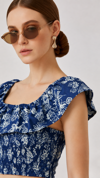 A close-up of a woman wearing an EVERSHE Prussian blue and white floral print ruffled top, gold-framed sunglasses, and gold earrings against a light gray background.