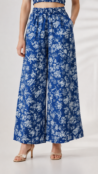 A model wearing blue wide-leg pants with a white floral print, paired with a matching blue crop top and beige heeled sandals, shown from the waist down against a light studio background.