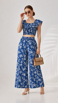 A woman models a matching two-piece set featuring a blue and white floral print, consisting of a ruffled crop top and wide-leg pants, styled with a brown handbag and sunglasses against a white studio background.
