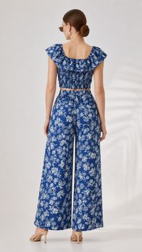 A woman shown from the back wearing a blue and white floral two-piece set with a smocked bodice, ruffled cap sleeves, and wide-leg pants, photographed against a light neutral studio background.
