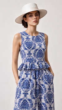 A woman wearing a sleeveless EVERSHE  white jumpsuit with a blue damask pattern and a matching white wide-brim hat, photographed against a light gray studio background.